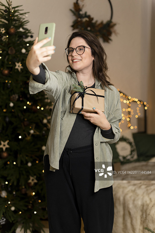 快乐的女人在家里用智能手机拍摄与圣诞礼物的自拍照图片素材