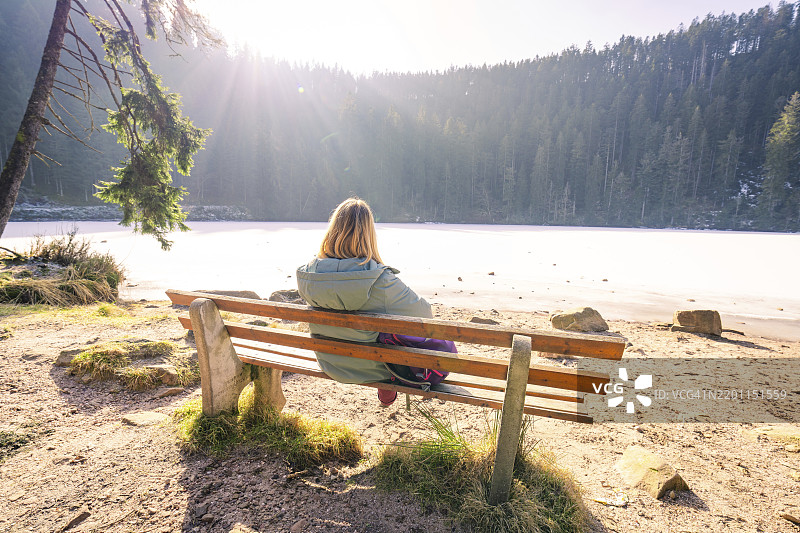 坐在冰冻湖边长椅上的女人,周围是明亮的冬季森林,格拉斯瓦尔德湖,巴德·里波尔茨瑙-沙普巴赫,沃尔法赫区,黑森林,德国,欧洲图片素材