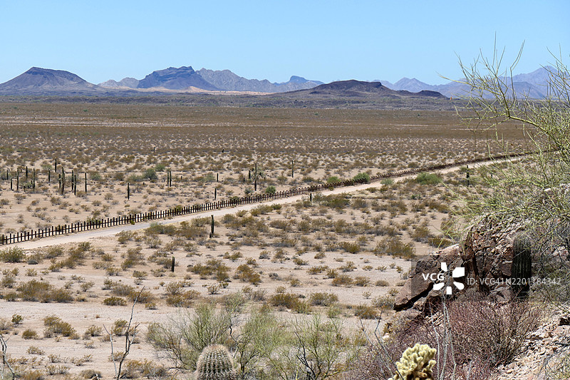 一张宽幅照片,展示了美国和墨西哥边界,由栅栏(墙)构成,背景是索诺兰沙漠中的仙人掌和山脉,位于亚利桑那州的卡贝萨·普列塔国家野生动物保护区。图片素材