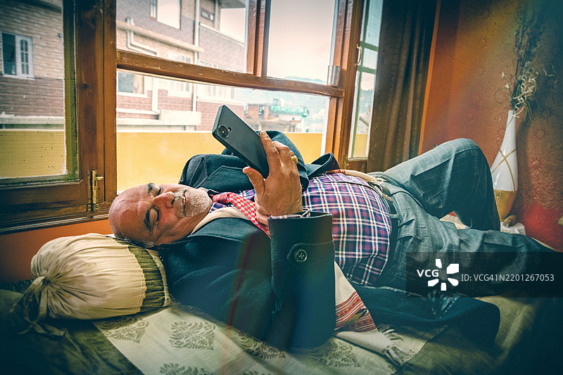 疲惫的老绅士穿着正式服装躺在床上看手机。图片素材