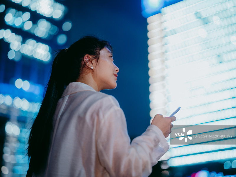 一位自信坚定的年轻亚洲女商人在金融区的现代摩天大楼前使用智能手机，仰望夜空。图片素材