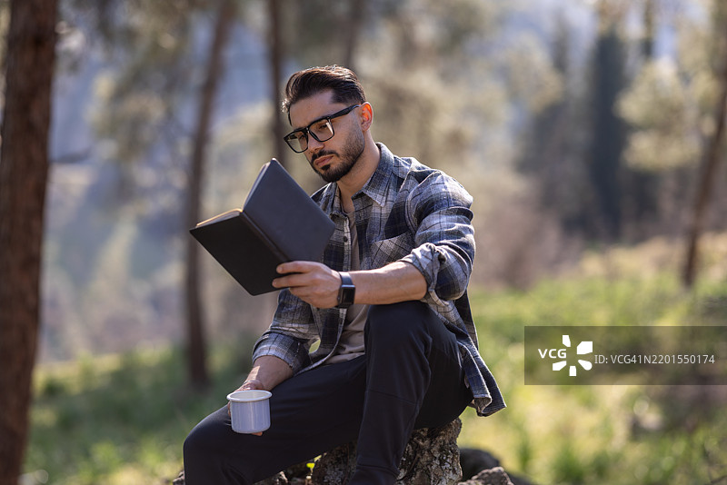 年轻男子在森林中享受宁静时光，阅读一本书并喝着咖啡图片素材