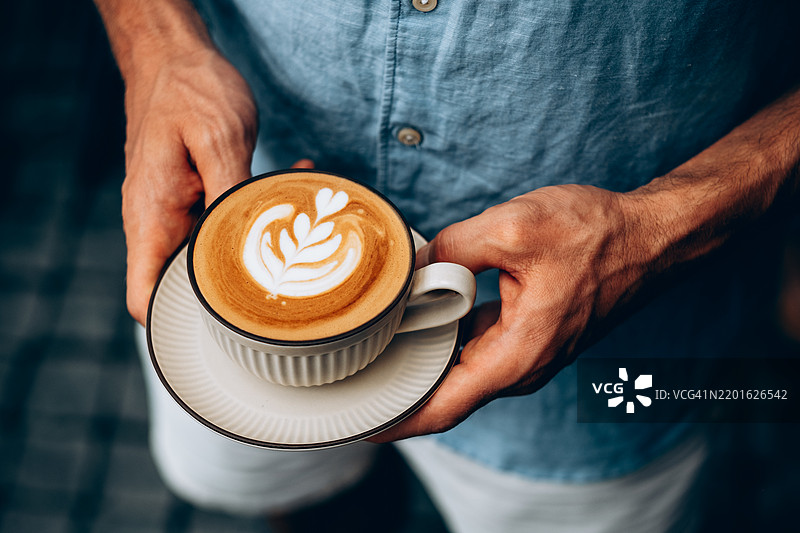 穿着蓝色亚麻衬衫的男子手握一杯带有拉花的卡布奇诺，双手轻轻托着碟子，突显出宁静放松的时刻。背景柔和模糊。图片素材