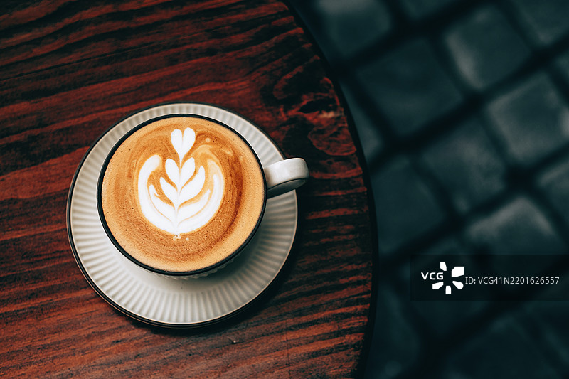 精美的卡布奇诺咖啡上有拉花，静静地放在木桌上。白色的泡沫图案与浓郁的棕色咖啡形成鲜明对比。图片素材