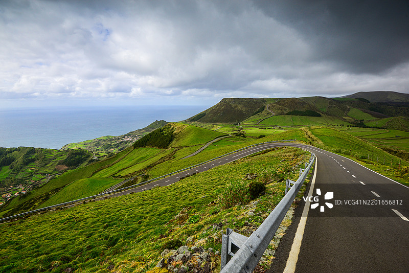一条蜿蜒的道路优雅地穿过生机勃勃的绿色山丘，通向在微云天空下壮观的海洋景色。图片素材