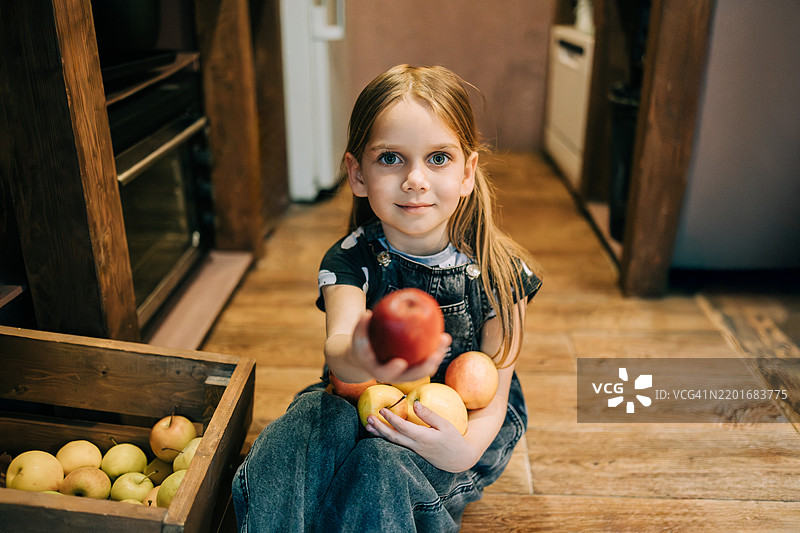 微笑的年轻女孩坐在厨房地板上，递出一个成熟的红苹果图片素材