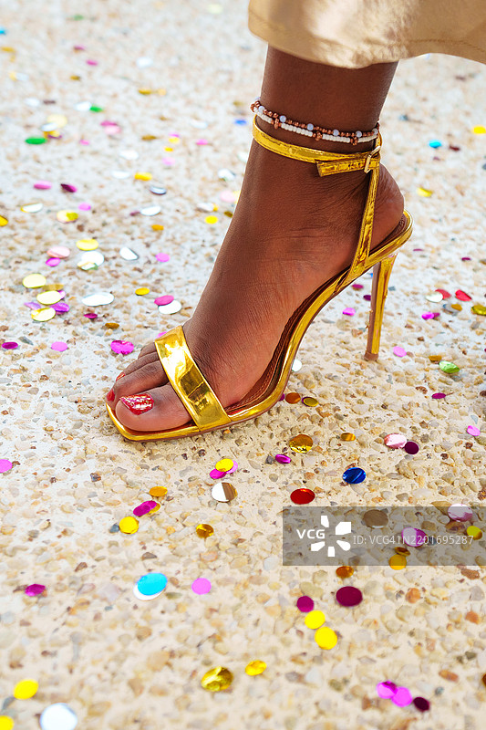 节日优雅与高跟鞋：女性优雅的腿部，佩戴手链，穿着时尚的金色凉鞋，站在派对五彩纸屑中。图片素材