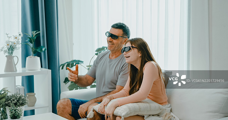 成熟的父亲和女儿在家中享受观看3D电影，佩戴着3D眼镜，坐在沙发上。家庭亲密和娱乐。图片素材