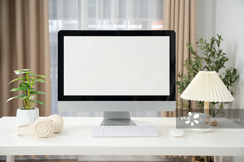 时尚工作空间的正面视图，配有电脑和办公用品。白色工作桌上的电脑显示器显示空白屏幕，旁边有无线鼠标和键盘，简约的办公室房间内装饰精美。图片素材