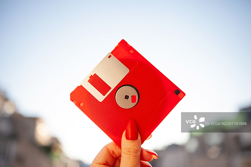 一只涂有红色指甲油的女性手握着一个红色软盘，背景是蓝天，两侧留有空白空间。图片素材