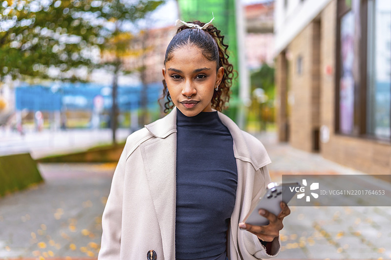 一位美丽的非裔美国女性在城市户外看着镜头，使用手机。图片素材