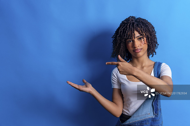 年轻女性用手指指向一侧，手掌张开展示某物，背景为鲜艳的蓝色。图片素材