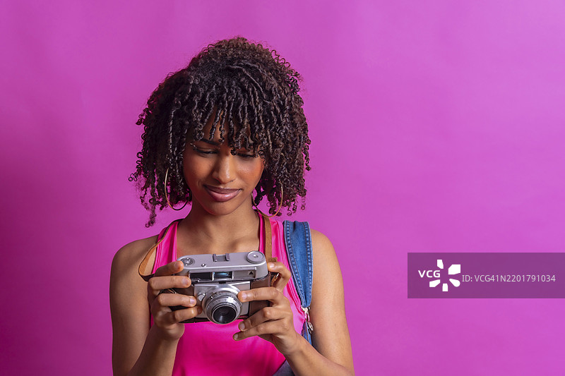 年轻女子背着背包，卷发，面对着她的复古相机，背景是鲜艳的粉色。图片素材