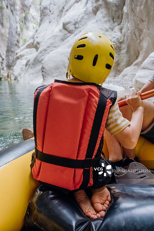 在风景如画的峡谷中，穿着安全装备的冒险漂流体验。一个穿着救生衣和头盔的孩子背影漂浮在漂流船上。图片素材