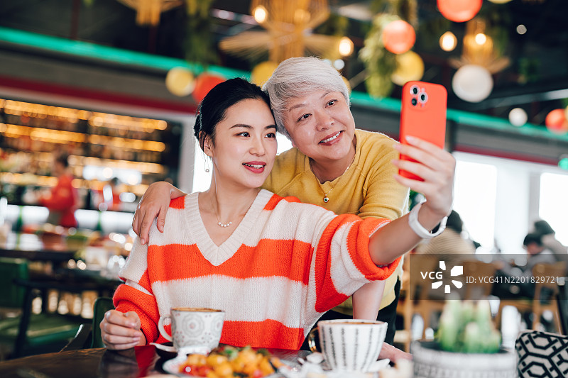 亚洲老年母亲和女儿在餐厅一起享用午餐，使用智能手机，享受美好时光。日常生活中的科技应用。人们、食物、生活方式与科技。图片素材
