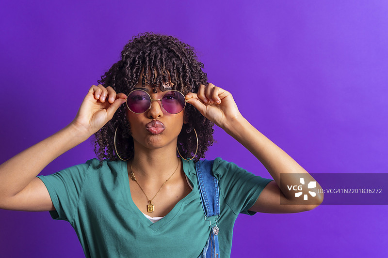 时尚的年轻女性，卷发，正在调整她的紫色太阳镜，并在鲜艳的紫色背景前撅嘴。图片素材