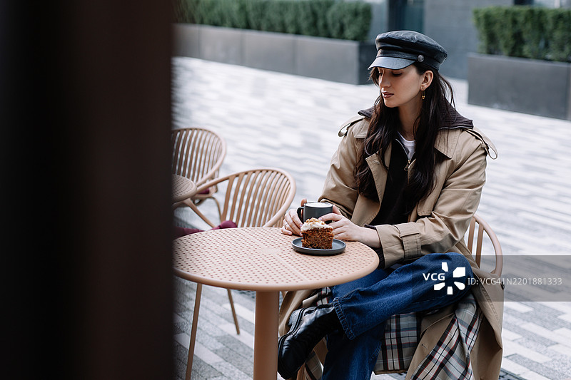 一位穿着休闲服和头饰的美丽女性坐在城市街道外的咖啡馆桌旁，喝着咖啡，吃着蛋糕。图片素材