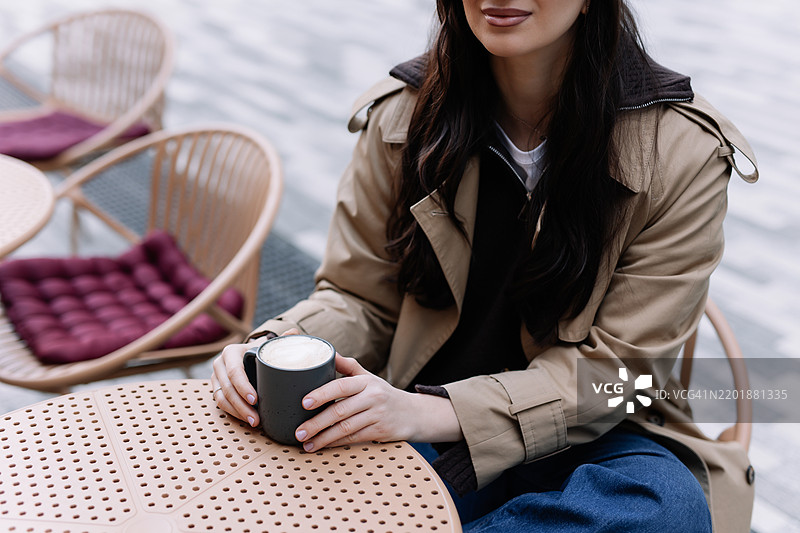 一位穿着休闲服和头饰的美丽女性坐在城市街道外的咖啡桌旁，喝着咖啡，吃着蛋糕。图片素材
