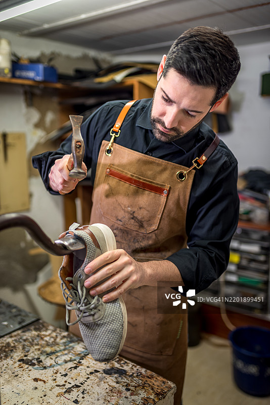 熟练的工匠在木制鞋撑上小心地锤打灰色运动鞋，细致地修理磨损的鞋子，工作在一个小型皮革作坊中。图片素材