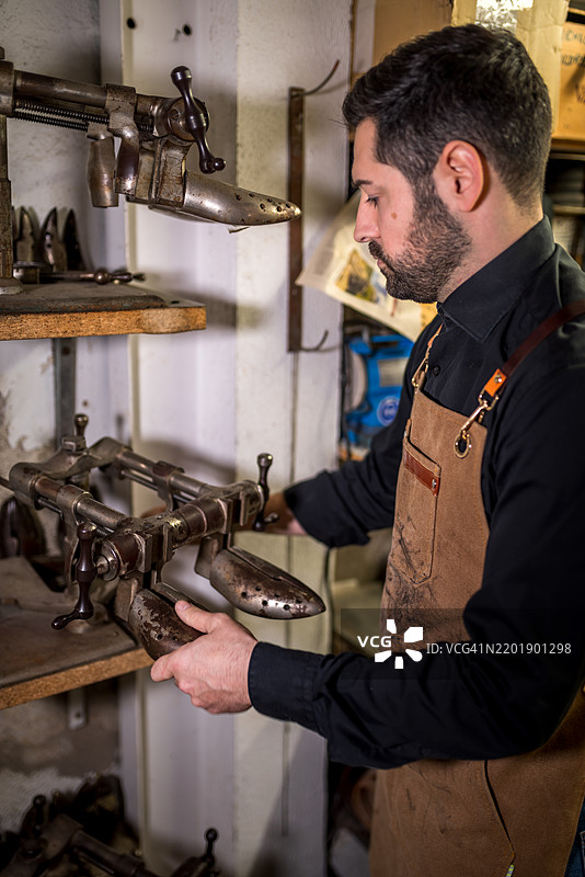 熟练的工匠在传统工作室的木架上挑选鞋撑，周围是复古的皮革工具。图片素材