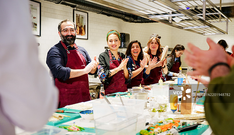 一位穿着白色制服的专业女厨师在为一群多元化的学生完成实践烹饪课程后，受到学生们的热烈 applauded。图片素材