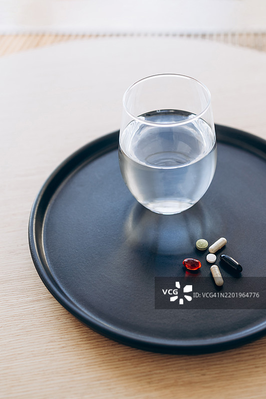 每日补充剂和水分。一杯水和各种维生素及补充剂整齐地摆放在黑色托盘上。健康、日常生活和自我护理的概念。图片素材
