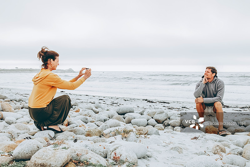 在宁静的海滩环境中捕捉快乐的亲密时刻。一位穿着黄色连帽衫和长裙的女性蹲在岩石海滩上，正在拍摄坐在海岸石头上的男性。图片素材