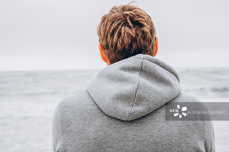 海边的孤独：在海岸氛围中反思的时刻。一个穿着灰色连帽衫的男人的背影。男人欣赏着海滩上美丽的日落。休闲、放松、旅游和冥想的概念。图片素材