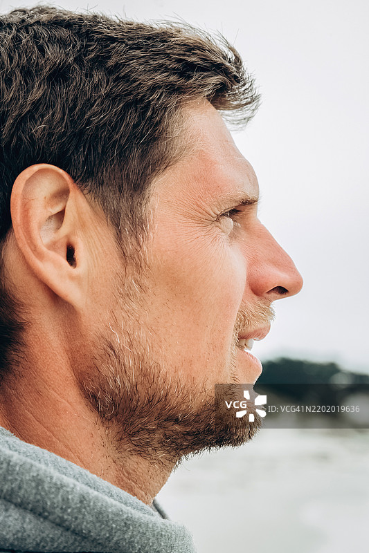 海滩上一个男人的沉思侧面肖像。身穿灰色连帽衫的帅气男人欣赏大自然，漫步在海滩上。男人欣赏海滩上美丽的日落。休闲、放松和生态旅游的概念。图片素材