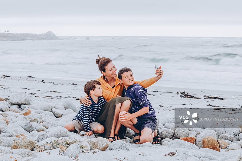 海滩上的家庭亲密时刻：母亲和两个儿子享受海边的快乐时光。母亲和她的两个儿子坐在岩石海滩上，微笑着一起自拍。图片素材