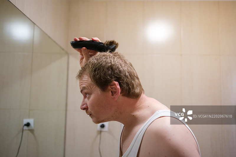 中年男子在浴室镜子前用电动剃须刀剃头。图片素材
