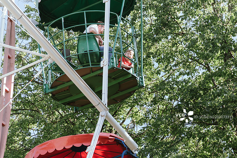 一个穿着红色T恤的小男孩在公园里和他的父亲一起坐摩天轮，显得非常开心。一起度过假期或周末的美好时光。图片素材