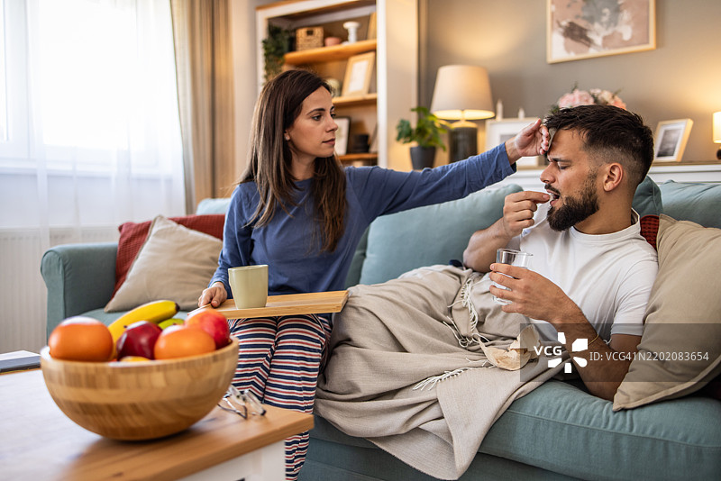 一位关心丈夫的妻子在客厅的沙发上照顾感冒的丈夫。图片素材
