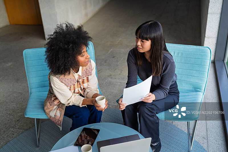 两位女商人在现代办公室的舒适椅子上讨论文件并喝咖啡。图片素材
