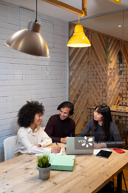 三位不同背景的同事在现代办公室空间中合作项目，使用笔记本电脑并做笔记。图片素材