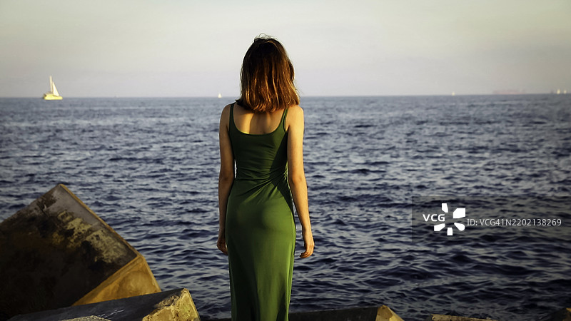 年轻女性背对着海洋，在日落时分靠近岩石欣赏风景图片素材