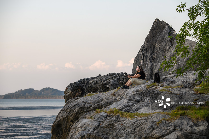 侧面视角的亚洲女性和博美犬坐在岩石上使用平板电脑图片素材