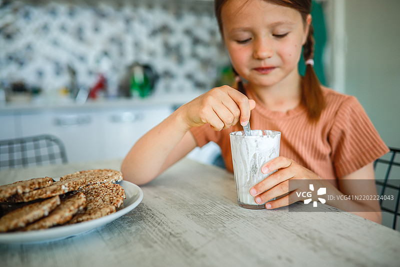 小女孩在厨房吃酸奶，忙碌的妈妈在背景中一边做多任务。图片素材
