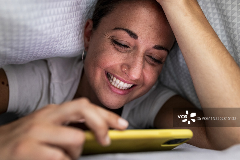 快乐的女人在家中用手机躺在毯子下图片素材