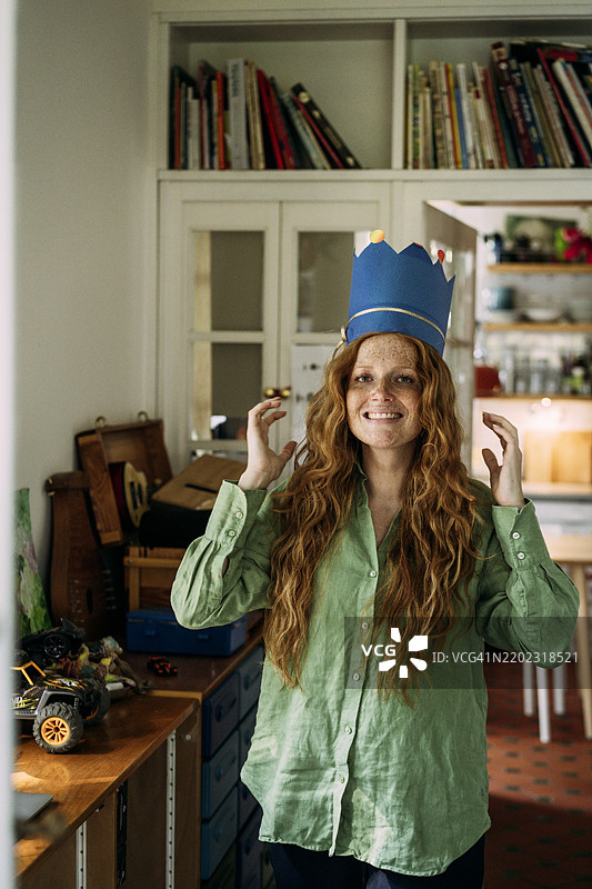 微笑的长发女性戴着纸制王冠帽，站在家中的卧室里图片素材