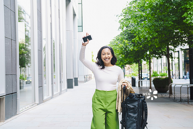 快乐的游客手持智能手机，拖着行李在城市中行走图片素材