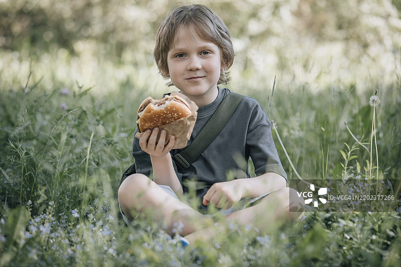微笑的男孩坐在草地上吃汉堡图片素材