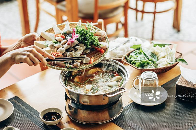 火锅餐桌上摆放着新鲜的蔬菜、海鲜和豆腐。一个人正在用筷子将食材放入沸腾的汤底中。桌子布置传统。图片素材