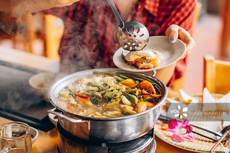 热气腾腾的海鲜汤锅，里面有虾、鱼和蔬菜，正在被端上桌。一位穿着红色格子衬衫的女士正在将海鲜舀到盘子里。图片素材