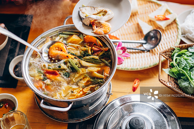 从上方俯视一锅热气腾腾的虾仁豆腐汤，汤中还有绿叶蔬菜。一个人正在添加新鲜食材。汤底浓郁芳香，周围摆放着海鲜和调料的盘子。图片素材