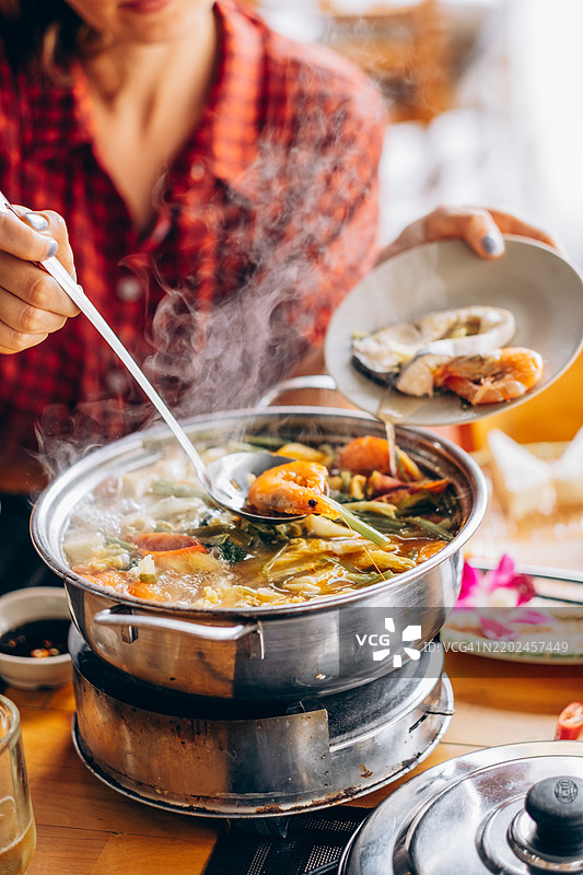 桌子上放着一锅热气腾腾的火锅，里面装满了虾、鱼、蔬菜和汤底。一个穿着红色格子衬衫的人正在用勺子舀虾，同时将更多海鲜放到盘子里。温暖的氛围暗示着一种舒适的用餐体验。图片素材