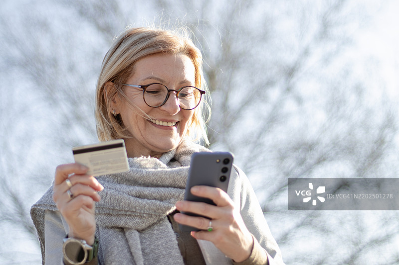 成熟的金发女性戴着眼镜，快乐地使用信用卡在阳光明媚的日子里网上购物。图片素材