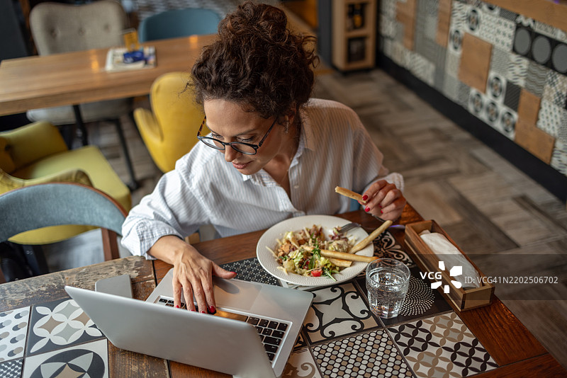 在餐厅里，一名自由职业者一边用笔记本电脑工作，一边吃沙拉。图片素材