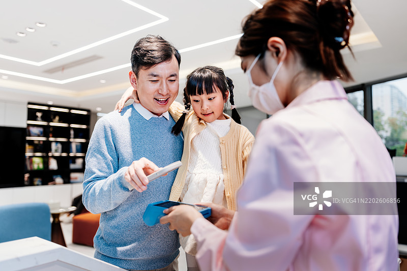 亚洲父亲在牙科诊所用智能手机付款，带着他的小女儿咨询牙齿问题。轻松的非接触式支付。NFC技术，轻触即付的概念。图片素材