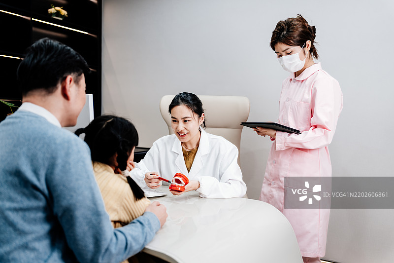 亚洲父亲带着他的小女儿在牙科诊所咨询。年轻的亚洲女医生手持牙齿模型，向小女孩解释牙科知识，提供友好的儿童牙科护理。图片素材